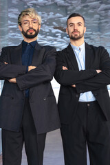 Vertical portrait Two confident male professionals in suits standing with crossed arms,serious at the camera inside a modern office. Corporate success, teamwork, and leadership in business environment