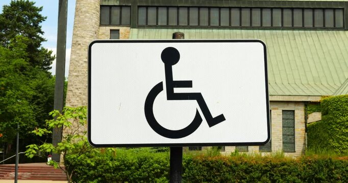White rectangular sign displaying black international symbol of access with person in wheelchair on street. Sign indicates accessible facilities for people