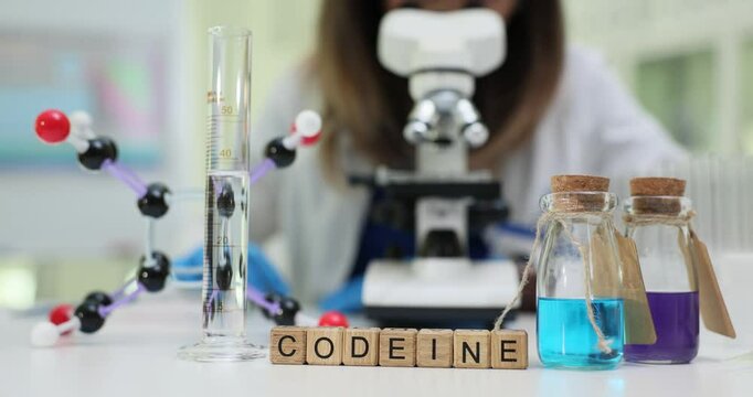 Word Codeine of wooden cubes and laboratory glassware near molecular structure model in lab. Woman researcher works with microscope in clinic