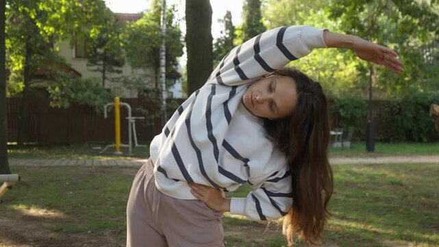 Woman performs a side bend stretch, warming up her body for an outdoor workout in a public park. Engaging in regular exercise promotes flexibility and a healthy active lifestyle.