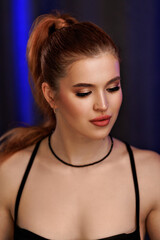 Close-up studio portrait of a woman with radiant bronze makeup and long ponytail, looking down with soft expression. Black outfit and gentle lighting create an elegant mood
