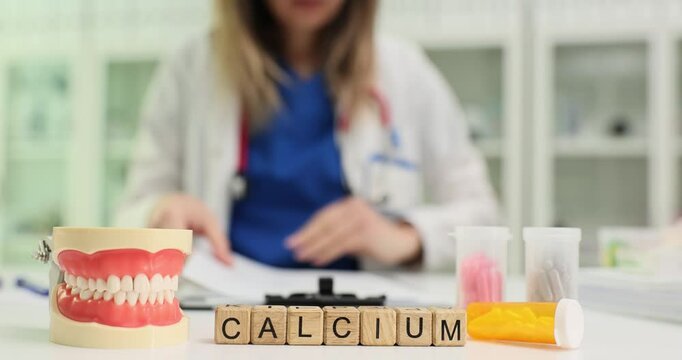 Calcium word on wooden blocks with human teeth model on doctor workplace. Woman physician recommends supplement maintaining strong bones slow motion