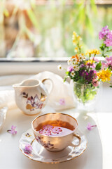 Creative postcard with porcelain cup of tea, teapot and a vibrant bouquet of garden flowers. Elegant tea set in beautiful soft sunlight. Good morning and teatime concept. Slow life visual trend.