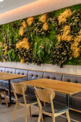 A cozy cafe space showcases a green vertical garden wall adorned with various plants. Stylish wooden tables and chairs offer a comfortable setting for patrons to enjoy their drinks