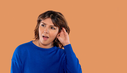 Surprised woman listening intently while holding her ear in a neutral setting with brown background...