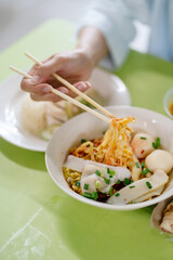 Hand holdind chopstick picking up a noodle from a bowl of dry noodles with assorted Asian street food surrounding on green table