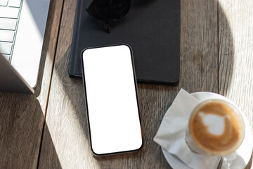 Smartphone with blank screen on wooden table next to laptop, notebook, sunglasses, and cappuccino. Cozy workspace setup perfect for remote work, freelancing, or creative productivity. Mockup phone