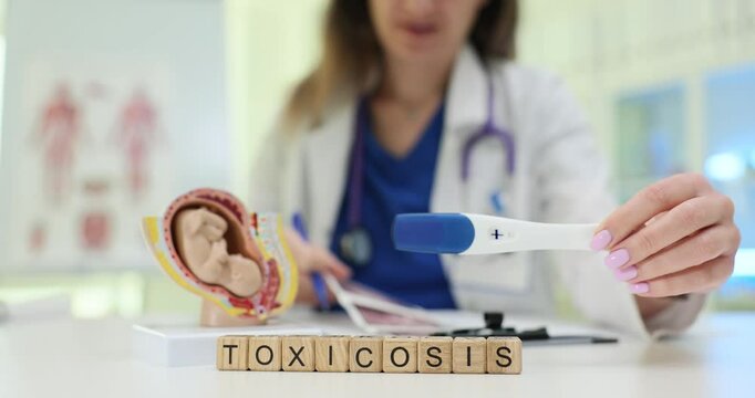 Text Toxicosis on blocks and gynecologist holding positive pregnancy test over desk. Doctor with anatomical model of fetus and ultrasound picture
