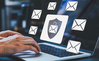 “A sleek digital illustration of email security featuring a hand typing on a silver laptop with floating translucent email icons and a central shield symbol against a dark, tech-inspired background