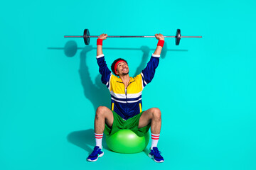 Young man lifting weights on exercise ball in retro activewear against vibrant turquoise background...