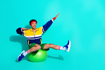 Fototapeta premium Young athletic man wearing retro sportswear having fun balancing on a fitness ball against a vibrant turquoise background