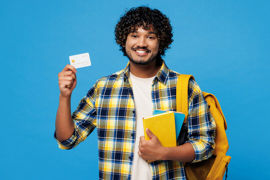 Young smiling happy fun Indian boy student wears yellow casual clothes backpack bag hold books credit bank card look camera isolated on plain blue background. High school university college concept.