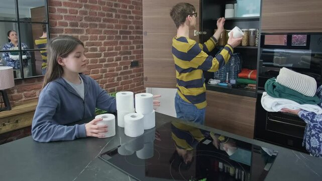 Brother and sister filling toilet paper while her mother storing clothes to stocked emergency food pantry in kitchen for natural disasters or war. Family preparing 72 hours survival kit in cupboard.