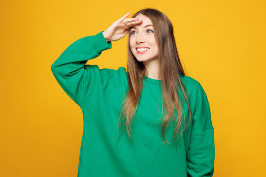 Young smiling happy Caucasian woman she wear green sweatshirt casual clothes hold hand at forehead look far away distance isolated on plain yellow orange background studio portrait. Lifestyle concept.
