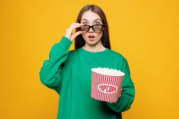 Fototapeta premium Young Caucasian woman she wears green sweatshirt casual clothes loer 3d glasses watch movie film hold bucket of popcorn isolated on plain yellow orange background studio portrait. Lifestyle concept.