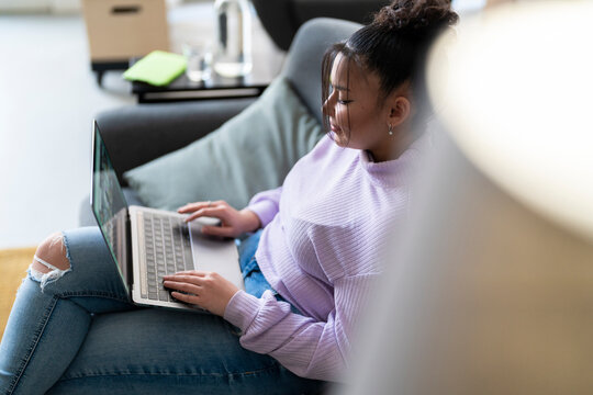 Young woman sits comfortably on a couch at home, working or studying on her laptop. She is focused on the screen, typing on the keyboard. Remote work or online learning concept.