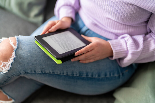 A woman wearing a purple sweater and ripped jeans is reading a book on an e-reader. She is sitting on a couch, enjoying her leisure time.