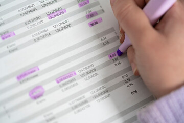 Close-up of a hand highlighting numerical data on a paper document using a purple marker. Focus on detail and analysis in business or finance.