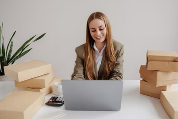 Woman in business attire working on laptop surrounded by packages in a modern office setting during...