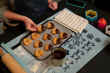 A person's hands dip heart-shaped cookies into melted chocolate on a baking sheet. Baking tools, cookie cutters, and a phone are on the table during a baking session.