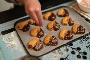A person's hand sprinkles colorful sprinkles onto heart-shaped cookies dipped in chocolate on a baking sheet. This is part of preparing sweet treats.