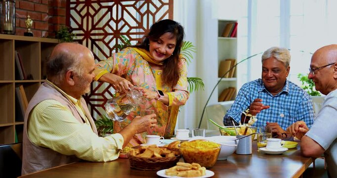 Seniors serving and eating together, Indian Asian old age friends reuniting at home eating food, serving snacks, cold drink, tea or coffee to each other while seated at modern dining table, enjoying