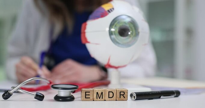Doctor reviews papers near wooden cubes with EMDR and anatomical eye model on desk. Psychological trauma therapy techniques at medical workspace