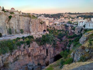 Dramatic landscape of Constantine, Algeria, during the golden hour light. It showcases the deep gorges carved by the Rhumel River, iconic limestone cliffs, the Bab Al-Qantara Bridge in the distance.