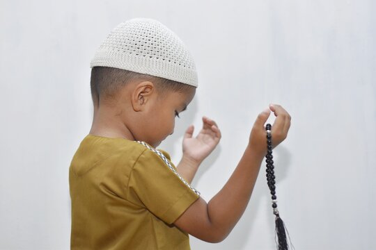 3 year old muslim boy raising hands in prayer while holding prayer beads, close up on white background