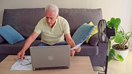 Video of Elderly man managing finances on a laptop while trying to stay cool using an electric fan - Powered by Adobe