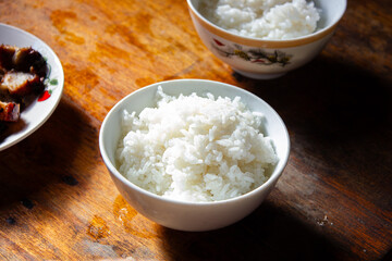 A close-up of a bowl of fluffy white steamed rice on a rustic wooden table, with another bowl and a plate of meat blurred in the background, evoking a comforting meal.

