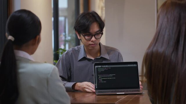 Programmer showing programming code on laptop screen to colleagues during informal team meeting, software engineer discussing project logic and development workflow with team members.