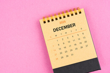 Brown desk calendar for December 2025 on pink background.