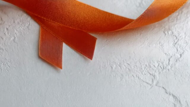 A shiny orange ribbon is diagonally placed on a white textured background, symbolizing support and awareness for a specific cause or medical condition.