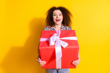 Smiling young woman holding large red gift box with bow against vibrant yellow background...