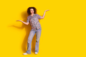 Young woman with curly hair gesturing positively, wearing casual printed shirt and jeans, standing in a cheerful yellow background