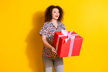 Joyful young woman holding a large gift box wrapped in vibrant wrapping paper on a yellow...