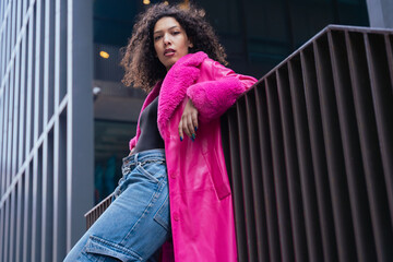 Graceful dancer with curly hair wearing a pink coat, expressing movement and energy in an urban environment. Modern fashion, contemporary dance, and artistic performance against a city backdrop.