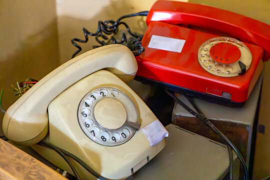 Two vintage rotary dial telephones — one beige and one red — placed side by side in a workshop or storage room. Classic devices from a bygone era with visible cords and markings. - Powered by Adobe