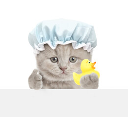 Cute kitten with shower cap on it head looks above blank white banner and shows thumbs up gesture and rubber duck. isolated on white background