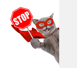 Happy cat wearing superhero costume looking from behind blank white banner and showing stop sign. Isolated on white background