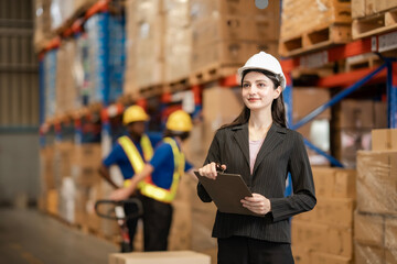 Confident female warehouse supervisor wearing a suit and hard hat, holding a clipboard, standing in front of stacked boxes and shelves. Logistics and supply chain concept.