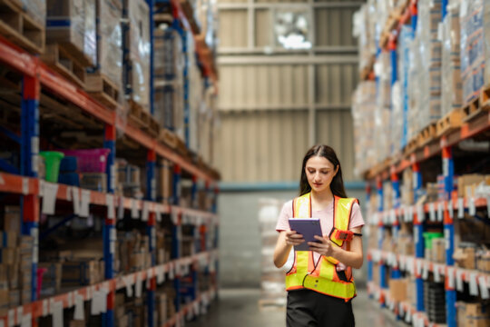 Warehouse employee wearing safety vest checking products with tablet. Female staff doing inventory in logistics center. Industrial workplace concept.