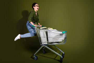 Fashionable mature woman joyfully posing with a shopping cart full of items against a vibrant green...