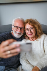 Happy senior couple taking selfie with smartphone while sitting on sofa in living room at home. Relationships and people concept
