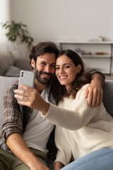 Happy young couple taking selfie with smartphone while sitting on sofa in living room at home. Relationships and people concept
