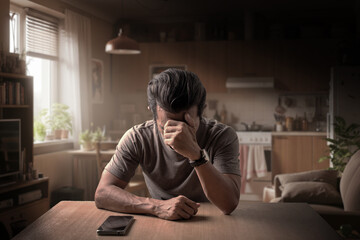 Fototapeta premium Sad man receiving bad news on phone, sitting at kitchen table in emotional distress, overwhelmed by grief and anxiety
