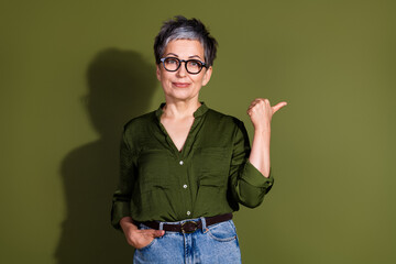 Confident mature woman in green shirt and glasses making a pointing gesture against a matching olive background