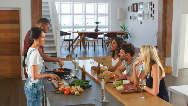 Couple Talking To Guests Whilst Making Food For Dinner Party At Home In Modern Kitchen