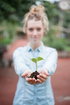 A young woman holds a small plant seedling in her hands, complete with soil. She extends her hands forward, presenting the new growth.